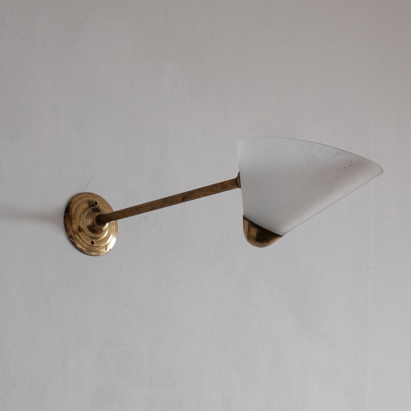 Wall-mounted light fixture with a cone-shaped white shade and brass arm on a plain background