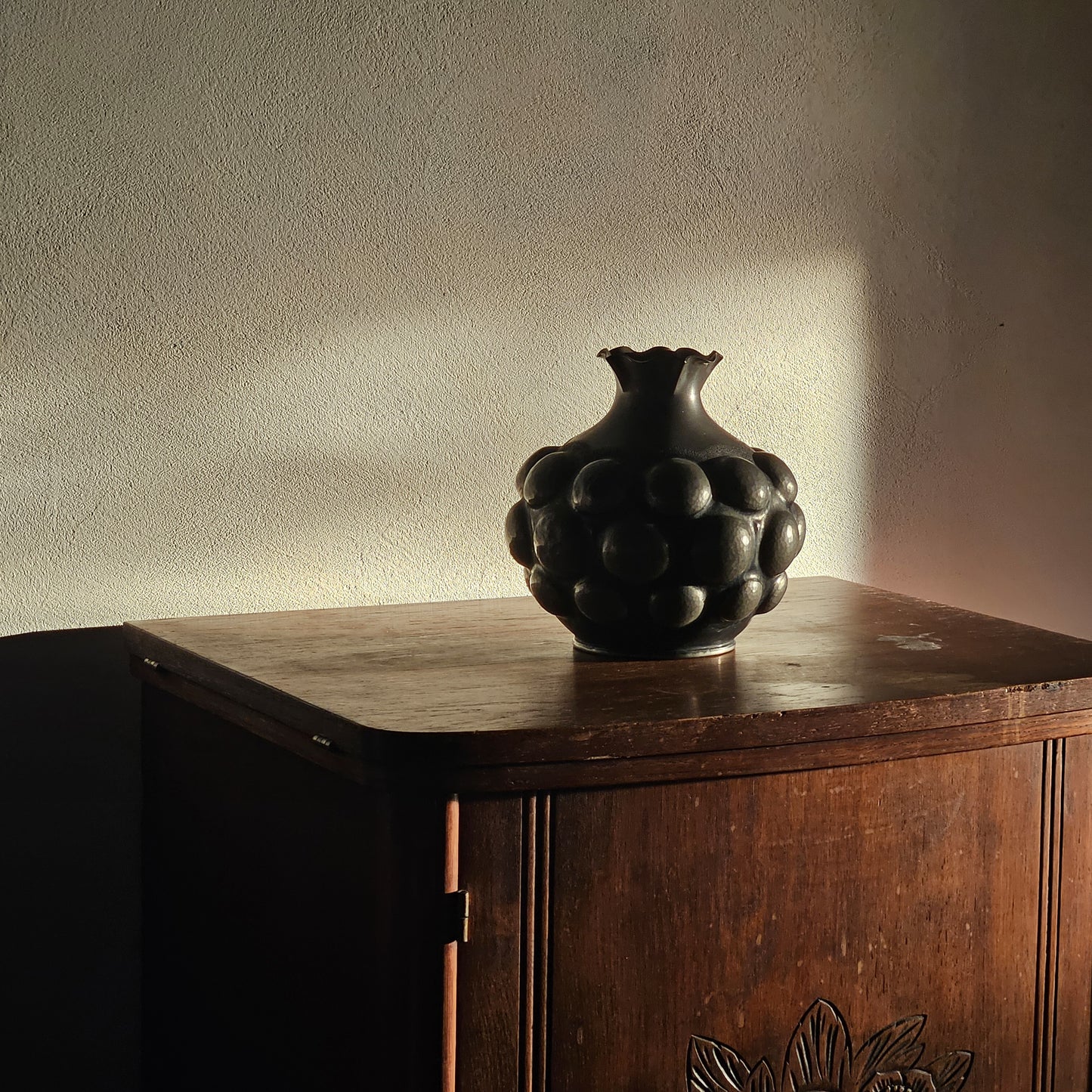Black decorative vase on a wooden cabinet against a textured wall.