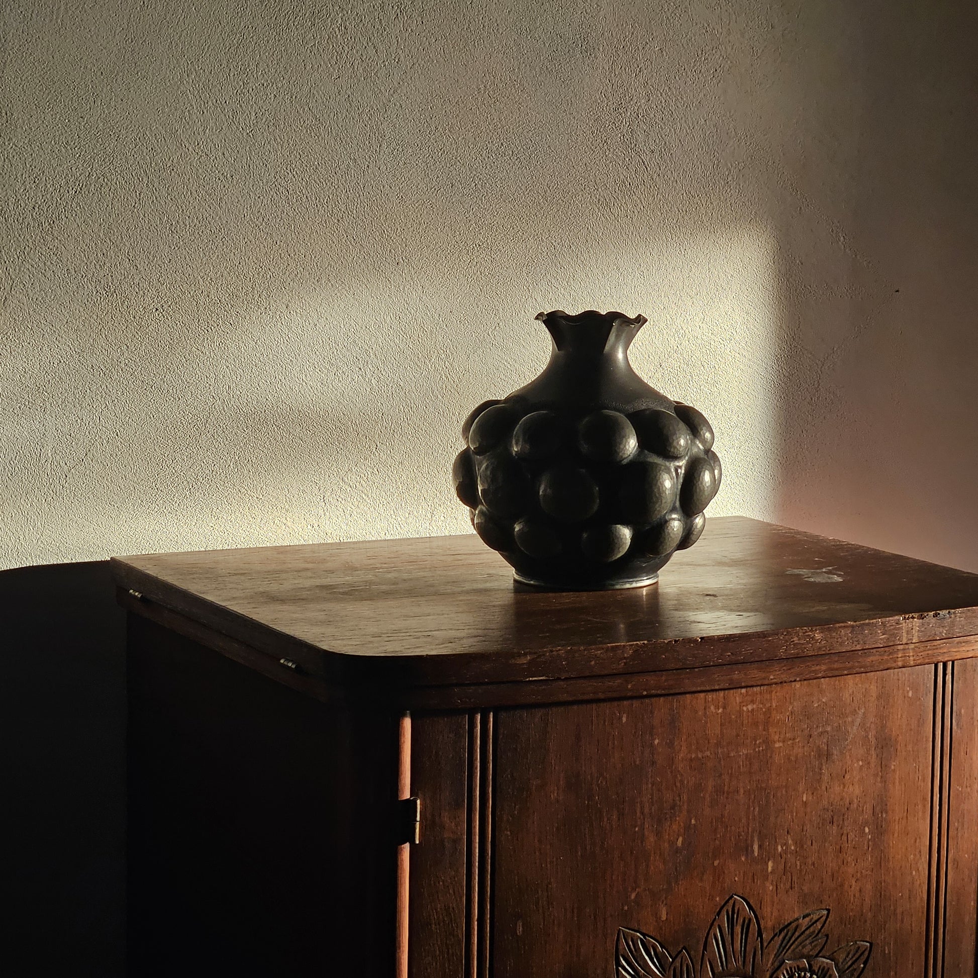 Black decorative vase on a wooden cabinet against a textured wall.