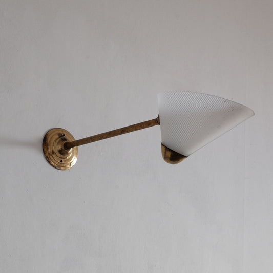 Wall-mounted light fixture with a cone-shaped white shade and brass arm on a plain background